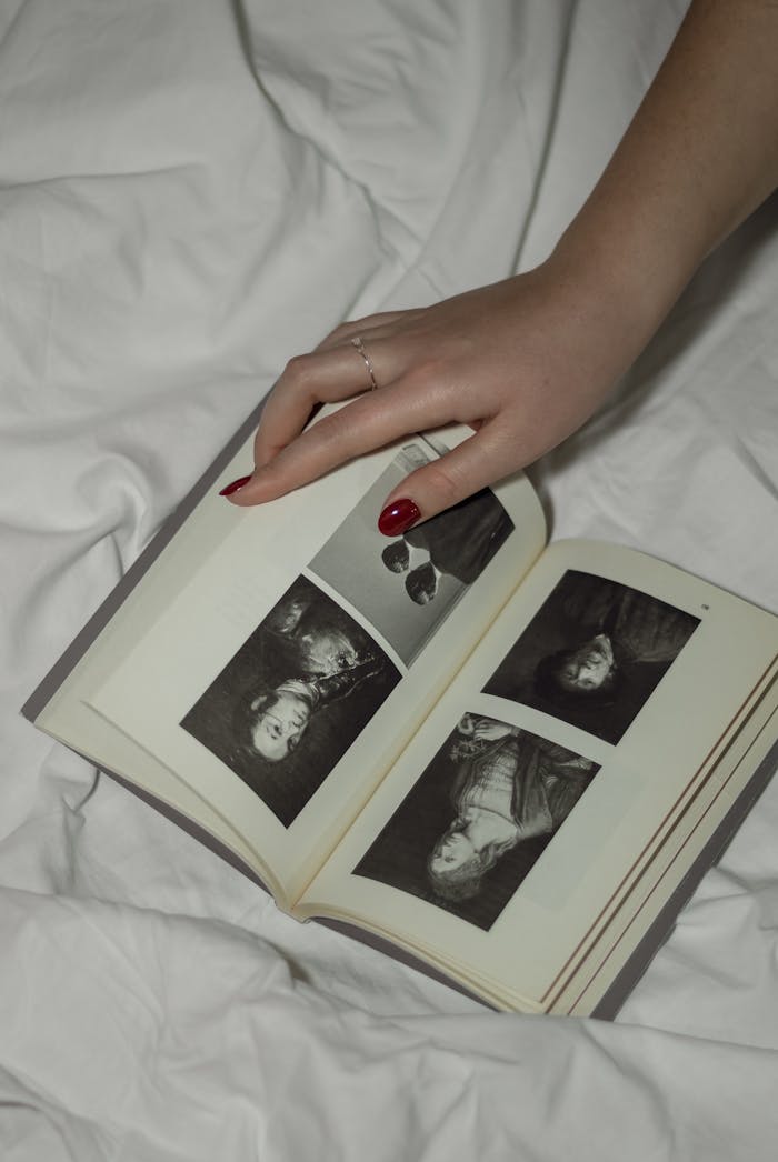 about-us-01 Close-up of a woman's hand holding a vintage photo album with monochrome portraits, resting on a white bed.