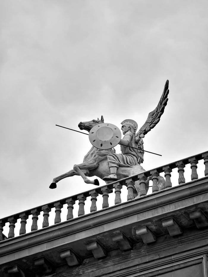 A dramatic black and white photo of a winged horseman statue atop an urban structure.